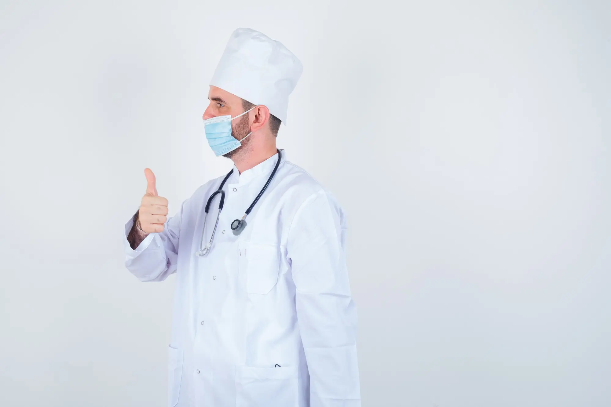Gutaussehender Mann mit um den Hals hängendem Stethoskop, der den Daumen nach oben zeigt, in weißem medizinischen Laborkittel und mit Maske, blickt selbstbewusst, Frontalansicht.
